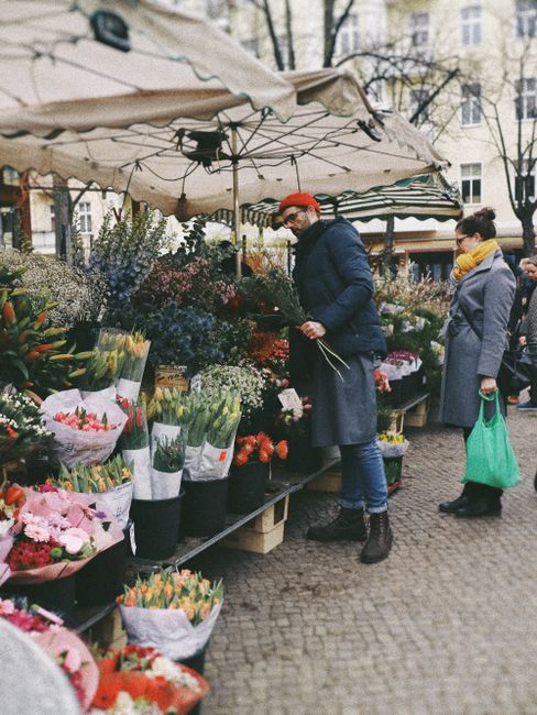 A woman and a man are shopping for fresh flowers at an outdoor market stall, which is covered by a beige canopy. The woman, dressed in a gray coat and yellow scarf, is holding a green reusable shoppin