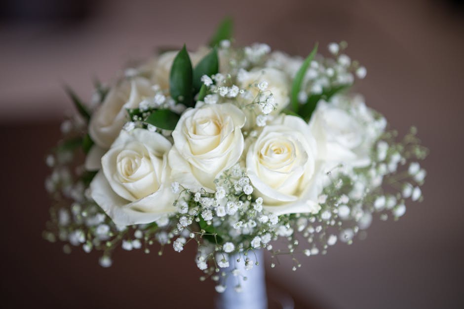 A floral bouquet featuring creamy white roses with fully open petals, accented by delicate sprigs of baby's breath and lush green leaves. The arrangement is placed in a clear glass vase, showing the f