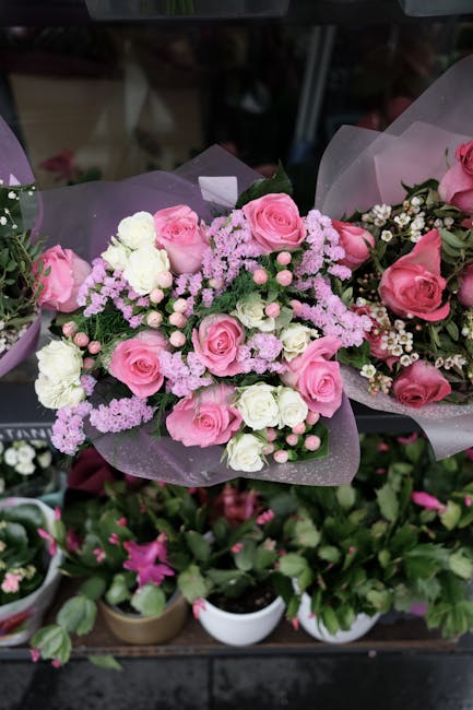 A bouquet of fresh pink roses and white spray roses, accented with small pink and white filler flowers, arranged in a rounded, lush floral display. The flowers are wrapped in delicate purple tissue pa