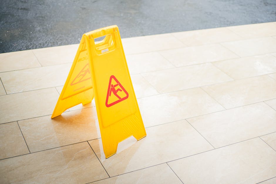 A yellow triangular 'wet floor' caution sign with red warning symbols stands on a beige tiled floor indoors, indicating a potentially slippery surface. The sign is positioned centrally, with natural l
