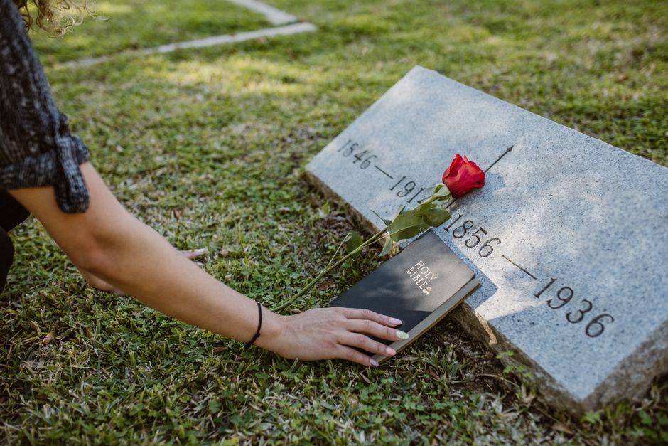 A person, wearing a short-sleeved top, is placing a red rose and a black Holy Bible on a grey stone grave marker that displays the years 1846-1956 and 1936. The person’s hand is gently resting on th