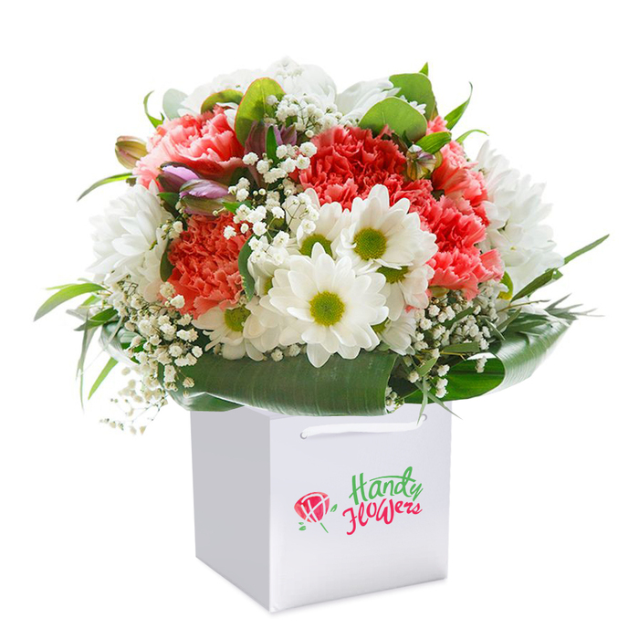 Arranged with artistry and a keen local eye, this elegant bouquet emerges from a pure white gift bag, echoing the contemporary yet homely style seen in Enfield Wash's favorite florists. The composition is anchored by plush coral and peach carnations, their plentiful ruffled petals adding richness and volume that catches both light and the observer's admiration. Interspersed among these focal blooms are perky white daisy chrysanthemums, each with a sparkling yellow-green center-bringing a lively, sunlit energy reminiscent of the vibrant community spaces near the Hertford Road market. Wisps of soft white baby's breath are threaded throughout, lending an ethereal, almost floating presence. Along the bouquet's edges and accentuating its round form, deep green, glossy foliage encircles the arrangement-a living frame that suggests growth and care. A handful of unopened buds with hints of purple and pink nestle quietly among the open flowers, hinting at the anticipation of visits or surprises yet to come. The bouquet is housed in a smart white gift bag with elegant ribbon handles, its minimal design highlighting the colorful blooms inside. Whether as a 'thank you,' a birthday wish, or a gesture of sympathy for someone along Ordnance Road, the arrangement radiates warmth, freshness, and genuine affection. Designed with the community's celebrations and quieter moments in mind, this display epitomizes the careful craftsmanship and considerate touch that Enfield Wash florists are known for.